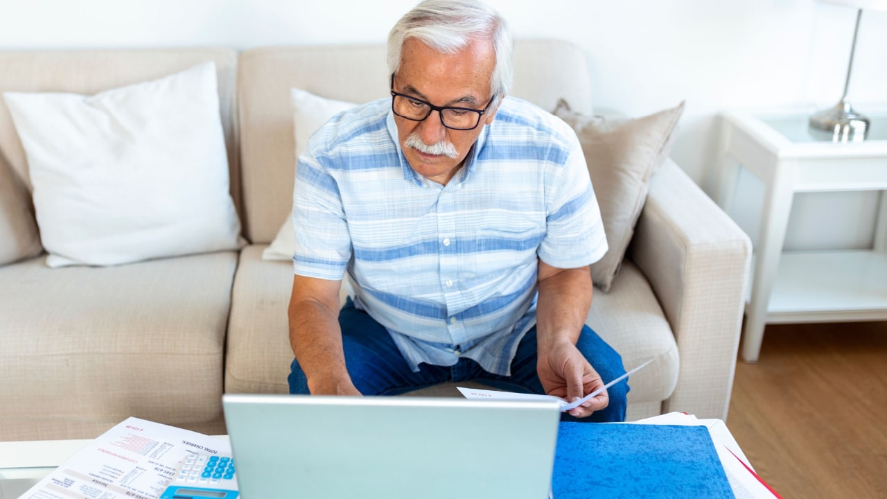Focused grey-haired elderly man sit on couch reading bank notifications calculating domestic expenditures, concentrated modern mature male consider financial paperwork, pay bills on laptop online