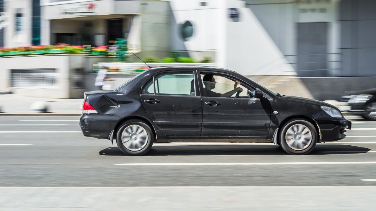 Moscow, Russia - July 2021: Mitsubishi Lancer sixth generation in black color running on the road with blurred background. Obsolete car with a dented rear fender in motion