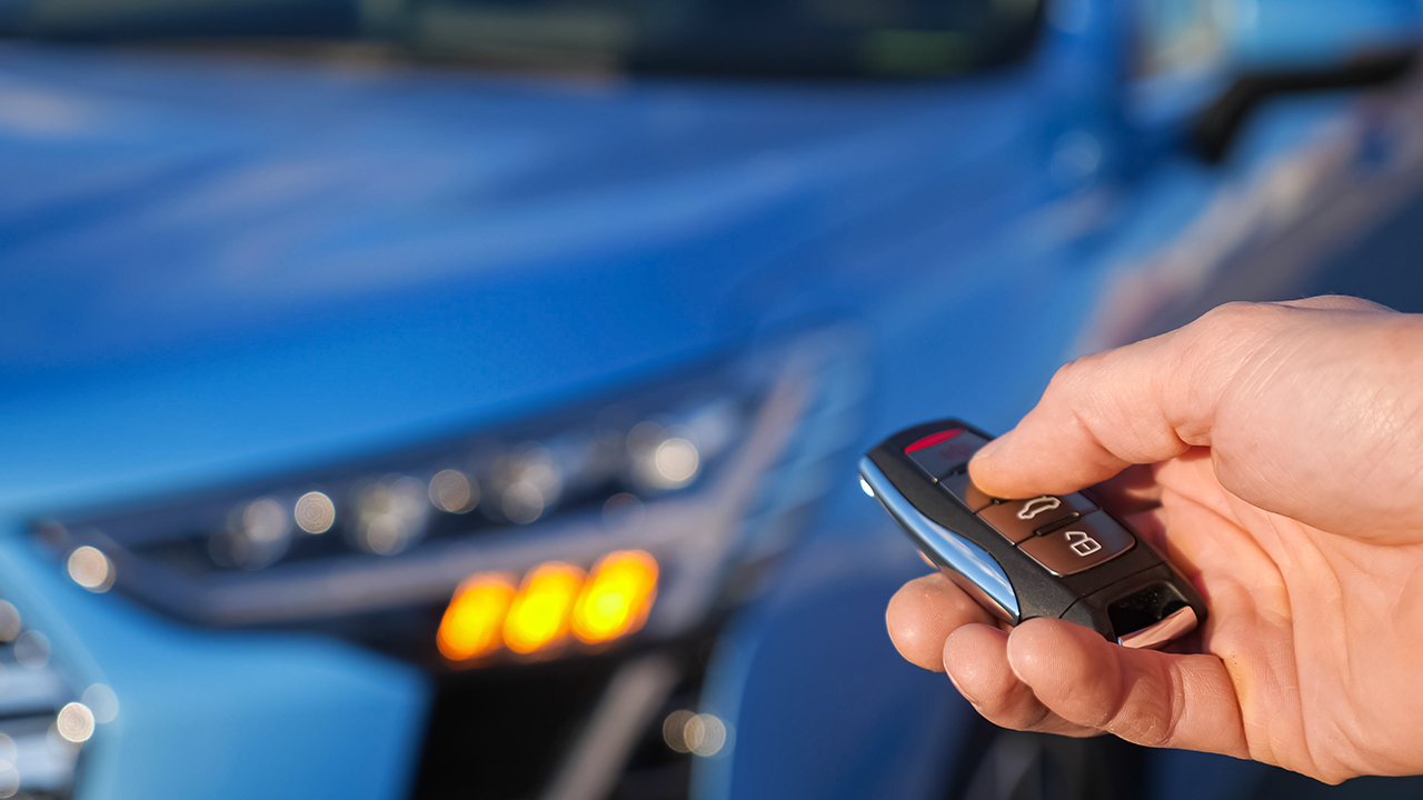 Man uses thumb to press black button on remote control key removing alarm signalling from bright blue car and headlights flash closeup